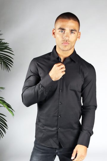 Vooraanzicht van heren overhemd in een zwarte kleur, met zwarte knoopjes en een slimfit pasvorm. Het model draagt een donkere jeans en heeft zijn kraag vast met zijn rechterhand