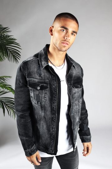 Vooraanzicht van model gekleed in zwarte denim jacket met beschadigde details en zilveren drukknopen. Het model heeft zijn armen losjes langs zich en kijkt recht in de camera.